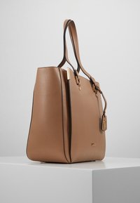 Torebka typu tote w kolorze brązowym z czarnym wykończeniem, złotymi detalami i dekoracyjną metką. Posiada usztywniony kształt oraz podwójne uchwyty.