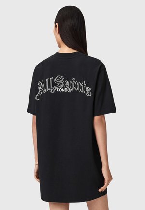 Kobieta z długimi ciemnymi włosami, ubrana w czarną, oversize'ową sukienkę-tunika z napisem "All Saints London" na plecach, stojąca na jednolitym tle.