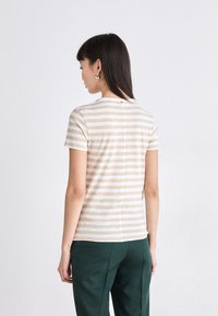 T-shirt rayé beige et crème à manches courtes, avec une couture arrière et un petit accent rouge au niveau du col. Associé à un pantalon vert.