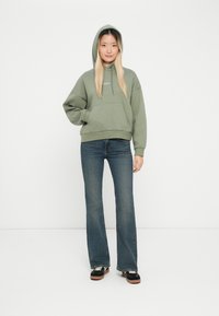 Olijfgroene hoodie met een kangoeroezak, gecombineerd met flared denim jeans, staande voor een effen witte achtergrond.