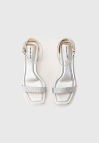 Silberne Absatzsandalen mit glänzendem Finish, einem glitzernden Riemen und Knöchelschnalle. Quadratische Zehenpartie. Markenname innen geprägt.