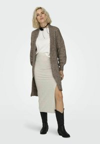 Larga chaqueta de punto marrón sobre una camiseta blanca, combinada con una falda gris clara con una abertura lateral y botas vaqueras negras.