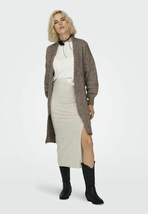 Larga chaqueta de punto marrón sobre una camiseta blanca, combinada con una falda gris clara con una abertura lateral y botas vaqueras negras.