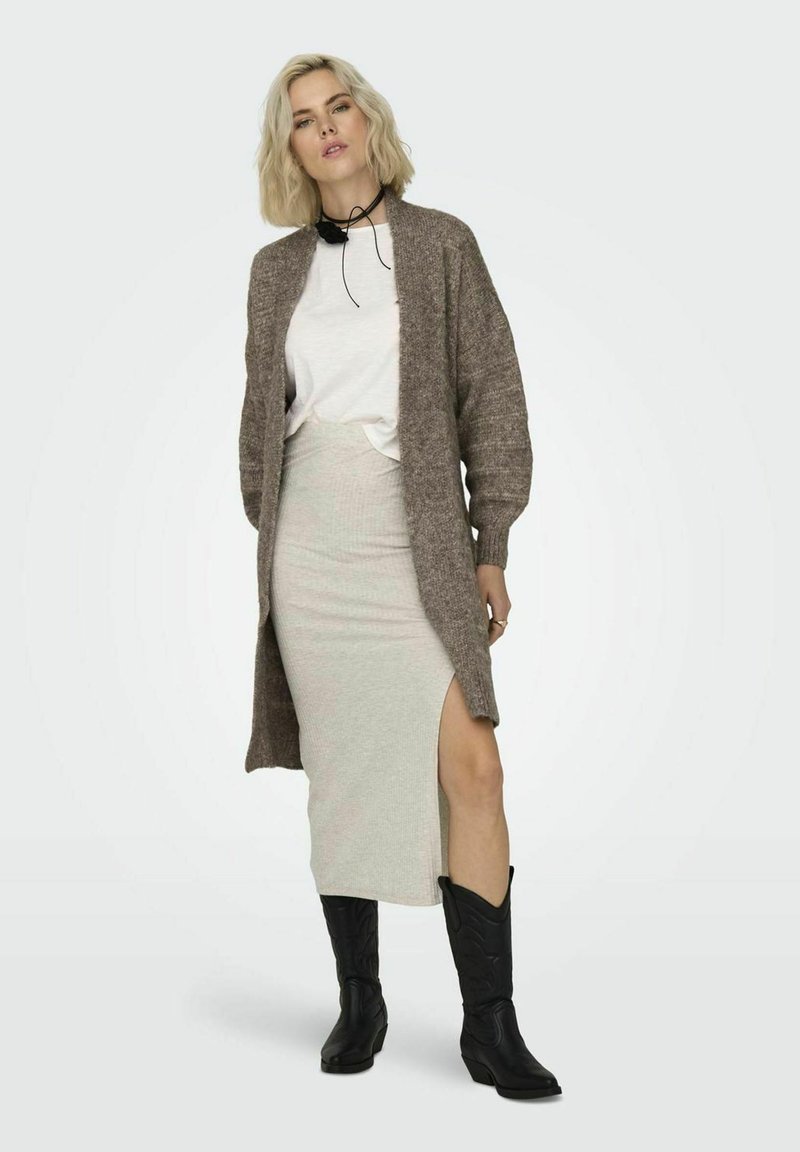 Larga chaqueta de punto marrón sobre una camiseta blanca, combinada con una falda gris clara con una abertura lateral y botas vaqueras negras.
