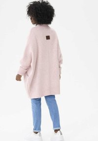 Różowy, oversize'owy sweter z dzianiny z wysokim kołnierzem, prążkowanymi mankietami i dołem, z brązową skórzaną metką z tyłu. Połączony z jasnoniebieskimi dżinsami.