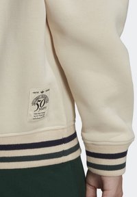 Felpa beige con maniche e orlo a righe blu navy e crema, dotata di un patch per il 50° anniversario e parte di un pantalone verde.