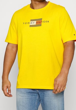 Yellow short-sleeve t-shirt with round neckline featuring a black and white striped logo "TOMMY HILFIGER" at the center. Soft texture.