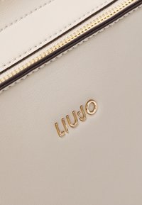 Detalle de un bolso de cuero crema con un cierre dorado y el logotipo en relieve "LIUːJO". Textura suave con costuras en contraste a lo largo de los bordes.
