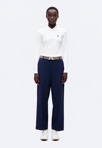 Femme debout, portant un polo blanc à manches longues, un pantalon large bleu marine avec une ceinture marron clair et des baskets blanches sur fond blanc.