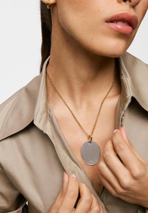 Mujer con collar de cadena de oro con colgante grande ovalado de plata, camisa beige con cuello y pequeño pendiente de aro dorado, sujetando parcialmente la camisa abierta.