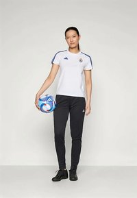 Noor naine seisab, hoides sinist ja valget jalgpalli, seljas valge Real Madridi särk, mustad Adidas püksid ja mustad jalanõud.