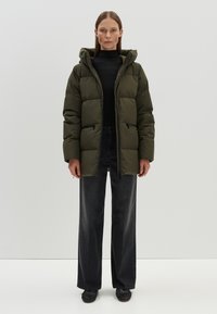 Olivgrüne Pufferjacke mit Kapuze, mit einem Reißverschluss vorne, zwei schwarzen Reißverschlusstaschen und elastischen Bündchen. Getragen über einem schwarzen gerippten Pullover und dunklen Jeans.