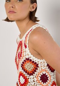 Virkat ärmlöst linne med ett blommönster i röd, orange, beige och vit. Har scalloped kanter och en texturerad yta.