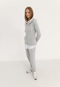 Felpa grigia con zip e tasche frontali, abbinata a pantaloni jogger coordinati. Realizzata in materiale morbido, indossata sopra una maglietta bianca, completata da sneakers.