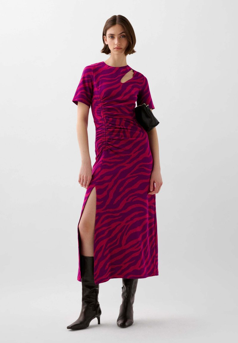 Fuchsia en donkerroze jurk met zebra-strepen, korte mouwen, een zijwaartse split, geplooide details en een uitsnijding bij de schouder. Gecombineerd met een zwarte tas en laarzen.