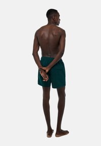 Shorts de bain texturés vert foncé avec une taille élastique et une poche arrière, présentés sur un modèle de dos, pieds nus et les mains jointes.
