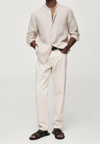 Homme portant une chemise beige à boutons, un pantalon beige clair, une ceinture noire et des sandales en cuir noir, debout les mains dans les poches.