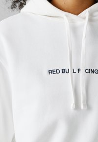 Persoon draagt een witte hoodie met trekkoorden en gedeeltelijk zichtbare zwarte geborduurde tekst met de woorden "RED BULL RACING" op de borst.