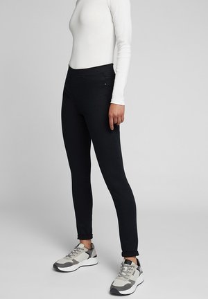 Stojąca osoba ubrana w czarne wąskie spodnie, biały top z długim rękawem i białe-szare trampki na jednolitym szarym tle.