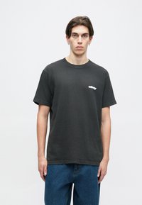 T-shirt de algodão preto com decote redondo e mangas curtas. Apresenta um logótipo branco no peito. Combinado com jeans de ganga azul.