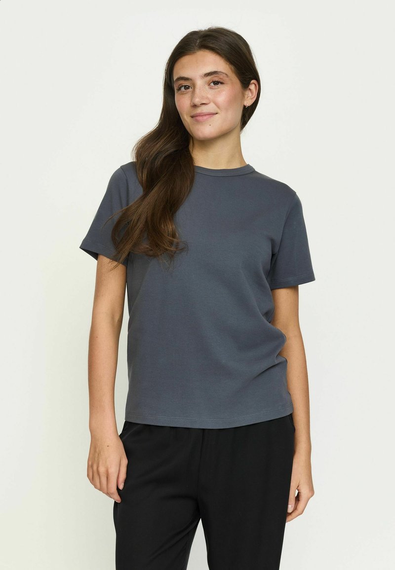 Femme aux longs cheveux bruns porte un t-shirt à manches courtes gris foncé et un pantalon noir, debout et souriante devant un fond clair et uni.