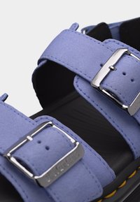 Κοντινό πλάνο λουριών σανδαλιών ανοιχτού μοβ χρώματος με ασημένιες μεταλλικές αγκράφες και μαύρη σόλα με λεπτομέρειες κίτρινου ραψίματος.