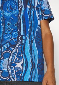 Närbild av en person som bär en kortärmad skjorta med blåa mönster, bestående av abstrakta och geometriska designer, med arm och del av torso synlig.