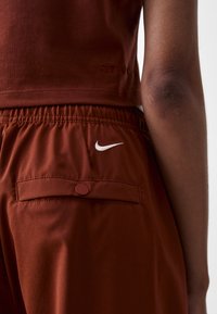 Rjav športni top z logotipom "DRI-FIT", v kombinaciji z rjavimi hlačami, ki imajo zadnji žep in bel Nike Swoosh logotip. Mehka tekstura tkanine.