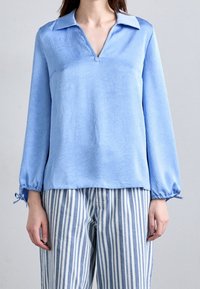 Světle modrá blůza s texturovaným povrchem, V-výstřihem a dlouhými rukávy; elastické manžety se stahovacími šňůrkami. V kombinaci s modro-bílými pruhovanými kalhotami.