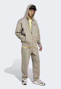 Beige urheilullinen verkkosuit, jossa on vetoketjuhuppari ja yhteensopivat housut. Ristikkäinen pinta logo-yksityiskohdilla. Täydennetty vaaleankeltaisella T-paidalla.