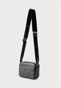 Μαύρη καπιτονέ τσάντα ώμου με ανάγλυφο μοτίβο MK και ρυθμιζόμενο υφασμάτινο λουρί κρεμασμένη πάνω σε απλό ανοιχτό φόντο.