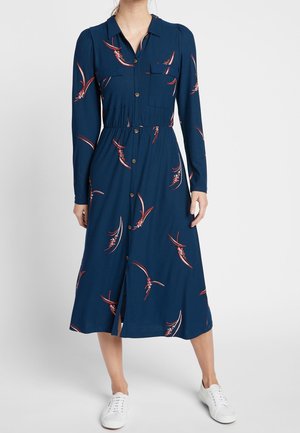 Kobieta ubrana w granatową sukienkę midi na guziki z czerwonymi kwiatowymi wzorami i długimi rękawami, w białych trampkach, stojąca na szarym tle.