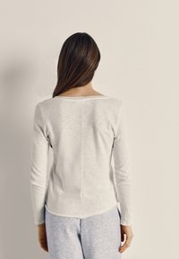 Pruunide pikkade juustega naine valges pika varrukaga särgis ja heledates hallides pükstes, seisab seljaga tavalise beeži värvi seina poole.