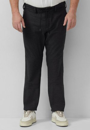 Mann mit dunkelgrauer Hose mit Kordelzug und Seitentaschen sowie weißen Turnschuhen, der vor einem einfarbigen hellgrauen Hintergrund steht.