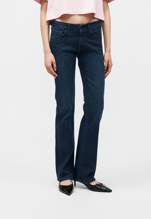 Γυναίκα που φοράει σκουρό μπλε τζιν με ίσια ποδόγυρα, ένα κομμένο ροζ τοπ και μαύρα ψηλά τακούνια με μυτερή μύτη και φιόγκους, στέκεται μπροστά σε απλό φόντο.