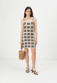Hihaton mekko, jossa on geometrinen musta ja kerma kuvio, ja neliöaiheita. Yhdistetty mustien hihnakenkien ja kudotun käsilaukun kanssa.
