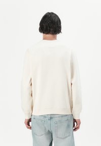 Person med krøllet mørkt hår, iført en cremefarvet sweatshirt og lyseblå jeans, vendt væk mod en ensfarvet hvid baggrund.