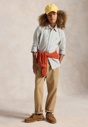 Enfant aux cheveux bouclés portant une casquette jaune, une chemise à rayures, un pull orange noué autour de la taille, un pantalon beige et des chaussures marron, debout les mains dans les poches.