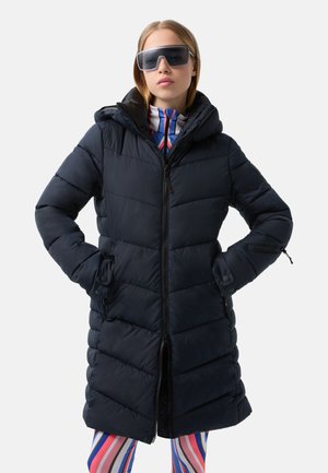 Navy puffer coat with a hood, horizontal quilted design, front zipper, and side pockets. Paired with brightly striped leggings.