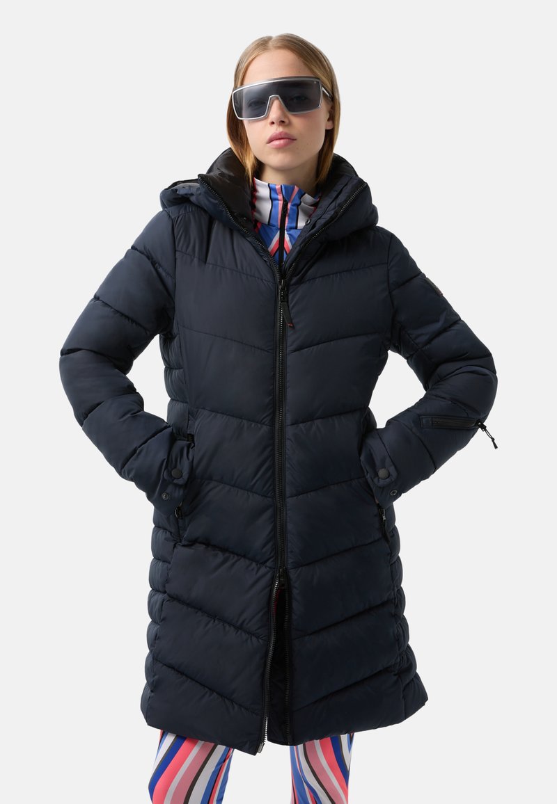 Navyblauer Puffermantel mit Kapuze, horizontal gestepptem Design, Frontreißverschluss und seitlichen Taschen. Kombiniert mit hell gestreiften Leggings.