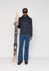 Sniega aprīkojums: tumši zils softshell žakete, zilas slēpošanas bikses, melni slēpošanas zābaki un rokās turēts rozā-melns slēpes ar stiprinājumiem. Uzvilkti tumši saulesbrilles.