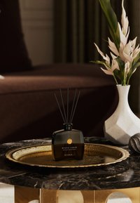 Czarny dyfuzor ze szkła z czarnymi patyczkami, oznaczony napisem "Mystic Cedar", umieszczony na złotej okrągłej tacy. Otoczony geometryczną wazą i ozdobnymi kamieniami.