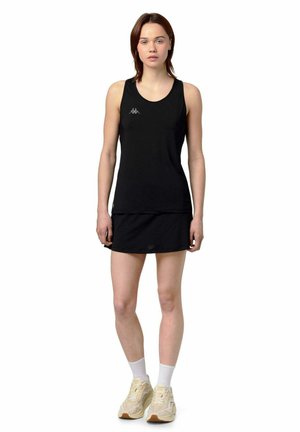 Jeune femme debout portant une robe de sport noire sans manches avec logo, chaussettes blanches et baskets beiges sur fond blanc.