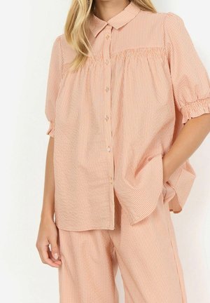 Woman wearing light orange and white striped button-up blouse with short puff sleeves and matching pants, one hand in pocket.