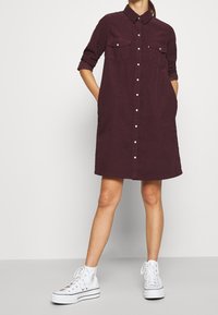 Robe-chemise bordeaux avec col, boutonnée à l'avant et deux poches sur la poitrine. Portée avec des baskets montantes blanches pour un look décontracté.