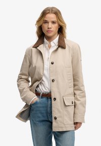 Mujer con chaqueta beige con cuello marrón, camisa blanca abotonada, vaqueros azules y cinturón trenzado marrón, de pie con una mano en el bolsillo.