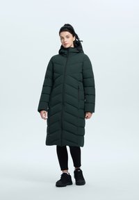 Abrigo puffer largo de color verde oscuro con costura en forma de chevron, cierre frontal con cremallera y capucha. Llevado sobre leggings y zapatos negros. Textura suave.