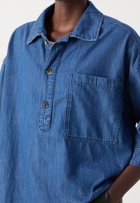 Denimhemd in Mittelblau mit Knopfleiste, spitzem Kragen, einer Brusttasche und sichtbaren Stichdetails. Weiche Textur.