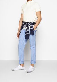 Bledě žluté polo tričko, světle modré kalhoty s ohrnutými manžetami a bílé tenisky. Námořnický pruhovaný svetr uvázaný v pase.