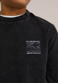 Schwarzer Fleece-Pullover mit rundem Halsausschnitt, strukturiertem Stoff und einem weißen Grafikdruck mit wellenförmigen Linien und Text auf der Brust.